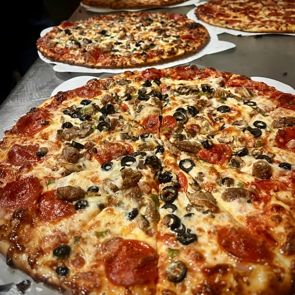 Multiple Task Force Pizza Co. pizzas, including one with pepperoni, olives, and mushrooms, on a metal counter.